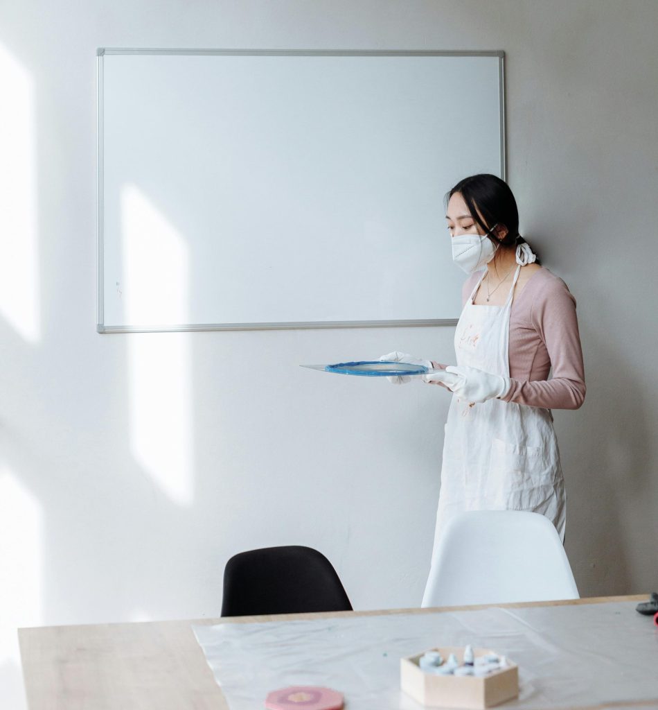 Asiatische Frau mit Schürze und Maske steht in einem Raum mit einer Tafel und hält Gegenstände in der Hand. Innenaufnahme.